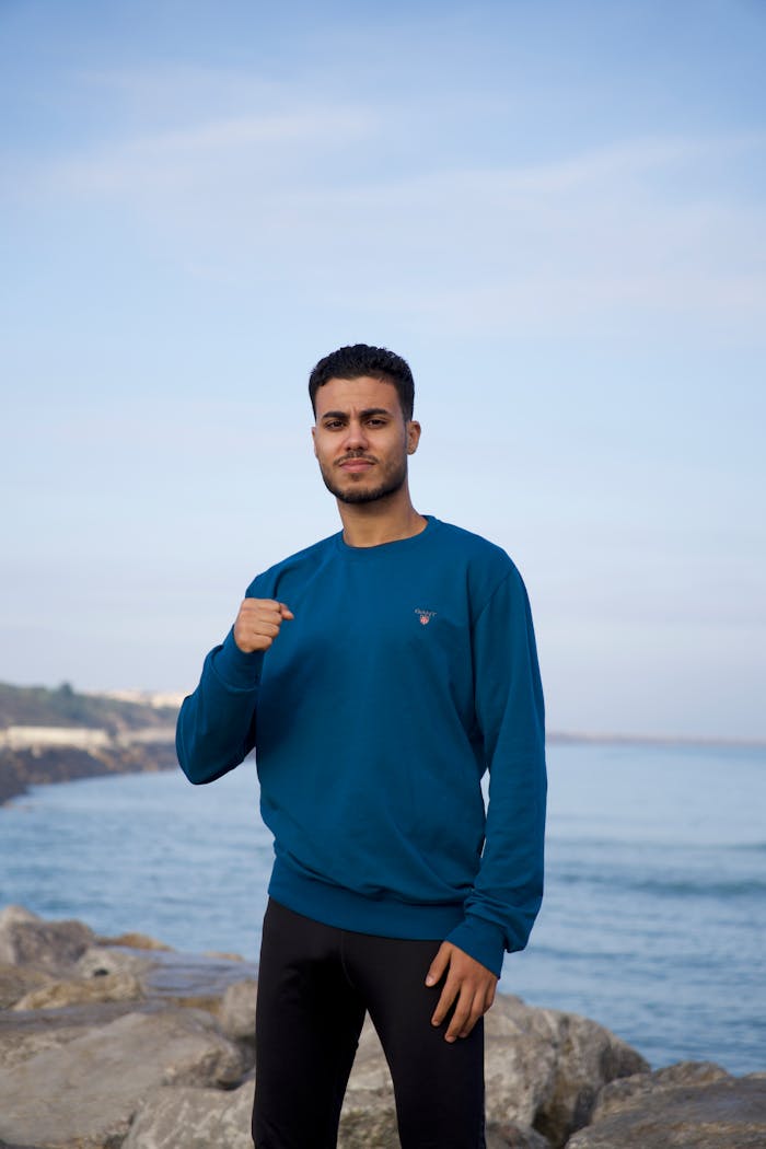 social-feed-img-04 Young man in blue sweatshirt posing on a rocky beach with ocean view.