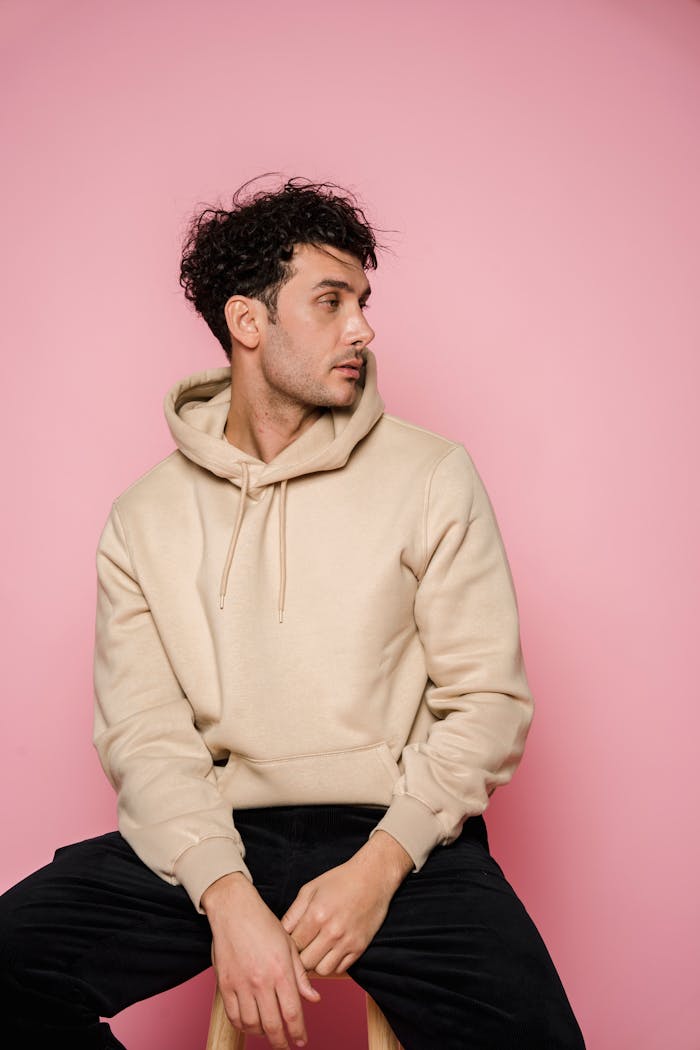quality-assurance-01 Thoughtful male with dark hair in stylish clothes sitting on wooden chair and looking away in studio against pink background